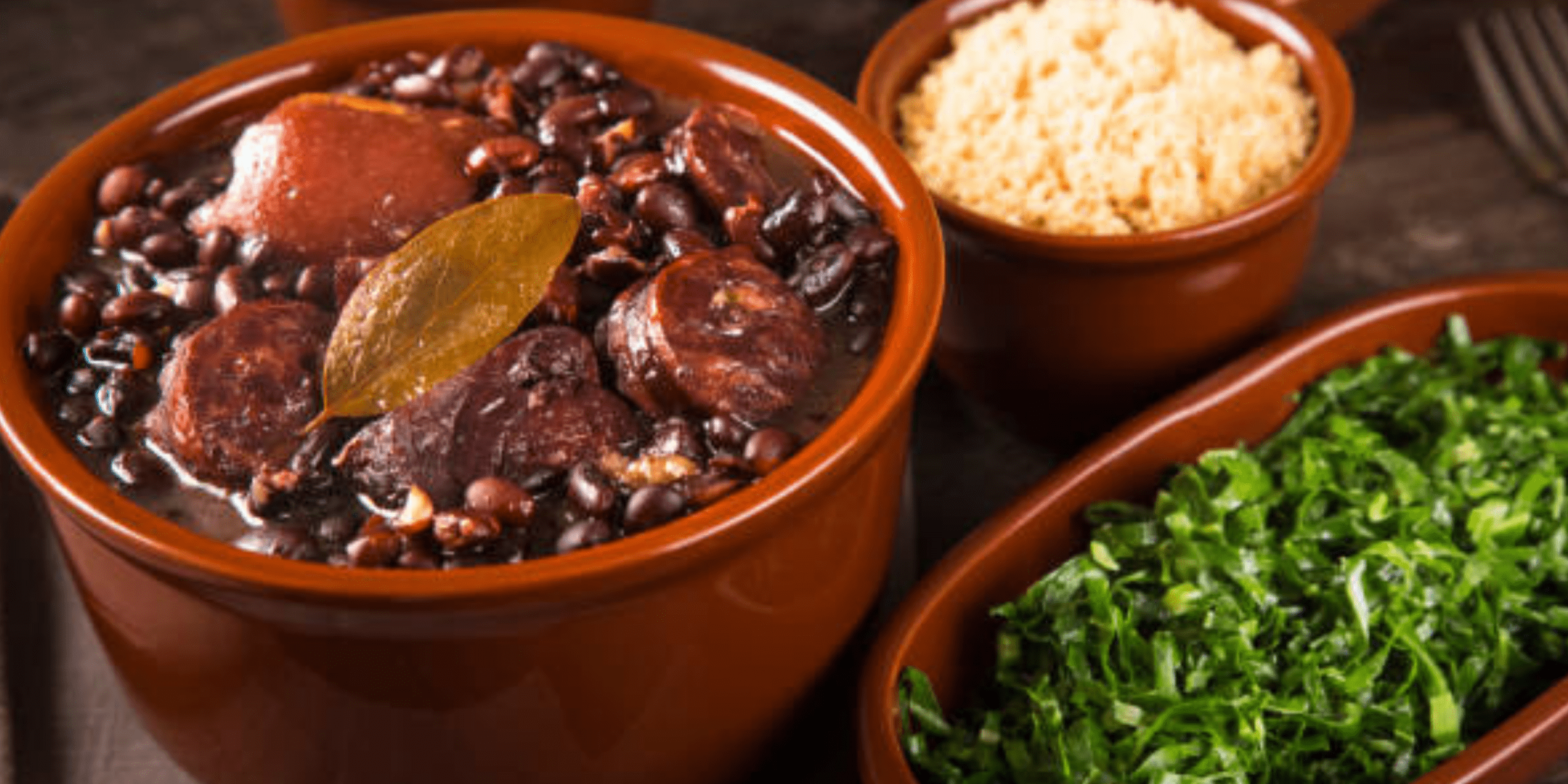 Feijoada servida em panela de barro, com feijão encorpado, carnes variadas como linguiça, carne seca e lombo, acompanhada de arroz branco, farofa e couve refogada em recipientes ao lado.