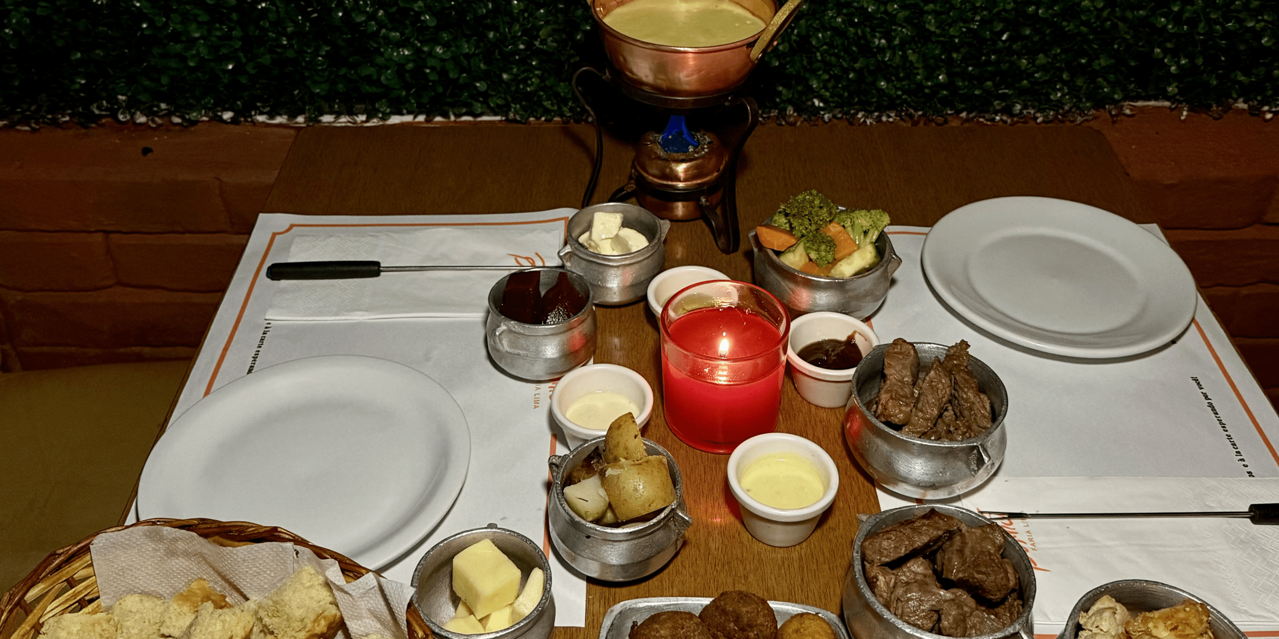 Mesa posta para fondue em ambiente aconchegante, com panela de queijo aquecida sobre rechaud ao centro, acompanhamentos variados como pães, carnes, legumes, batatas e molhos dispostos em pequenas panelas, além de vela acesa criando clima intimista para duas pessoas.
